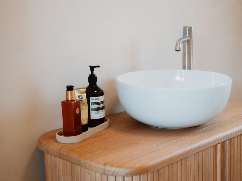 Lavabo a bacinella bianco con rubinetto alto in acciaio su mobile in legno con flaconi di detergenti